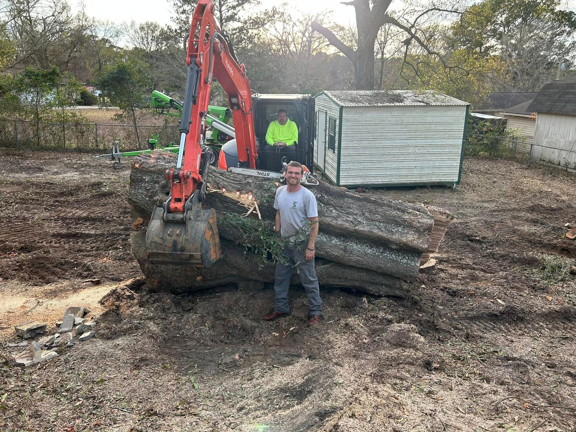 Man standing beside an orange excavator loading logs, with a metal shed in a muddy yard