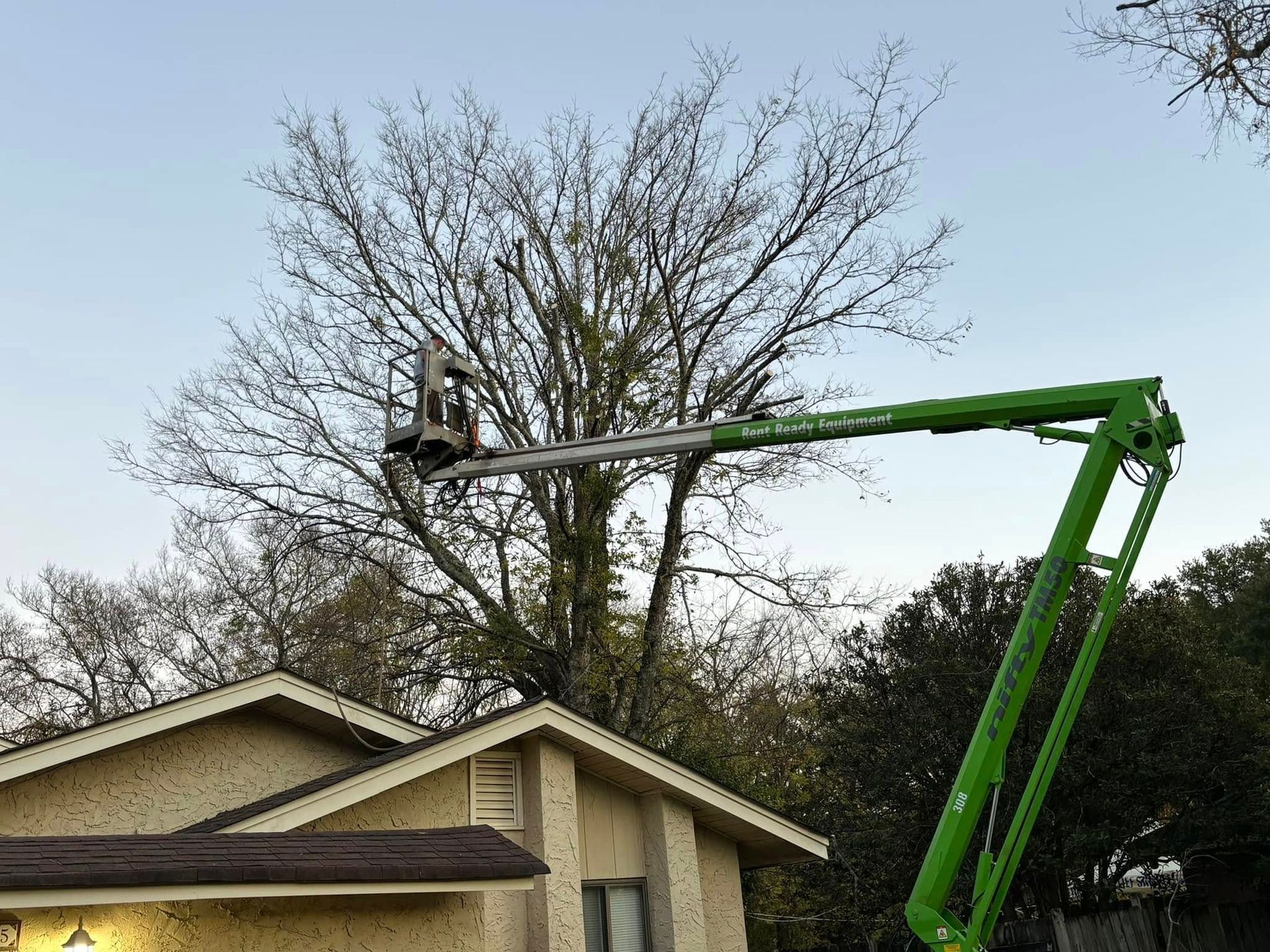 Worker in a green boom lift trimming tree branches above a house roofline