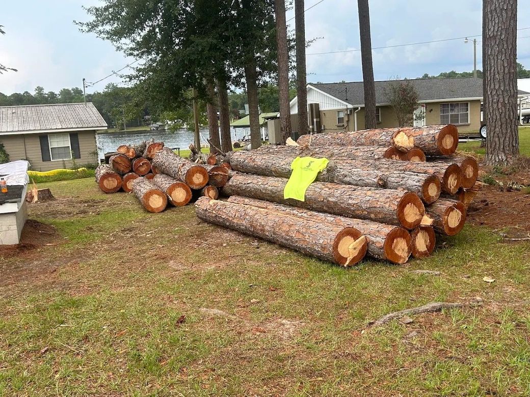 Stacked logs in a grassy yard near houses and pine trees, with a neon yellow tag on top.