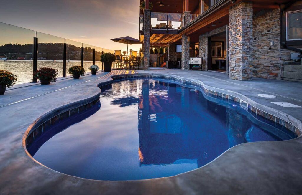 Curved blue pool beside a stone house at sunset, with a lake and mountains in the background