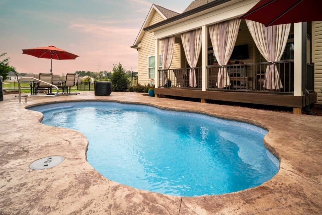Backyard pool beside a patio home with red umbrellas and lounge seating at sunset