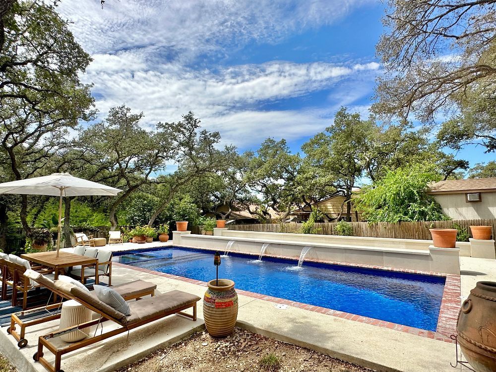 Backyard pool with lounge chairs, umbrella, and trees under a blue sky