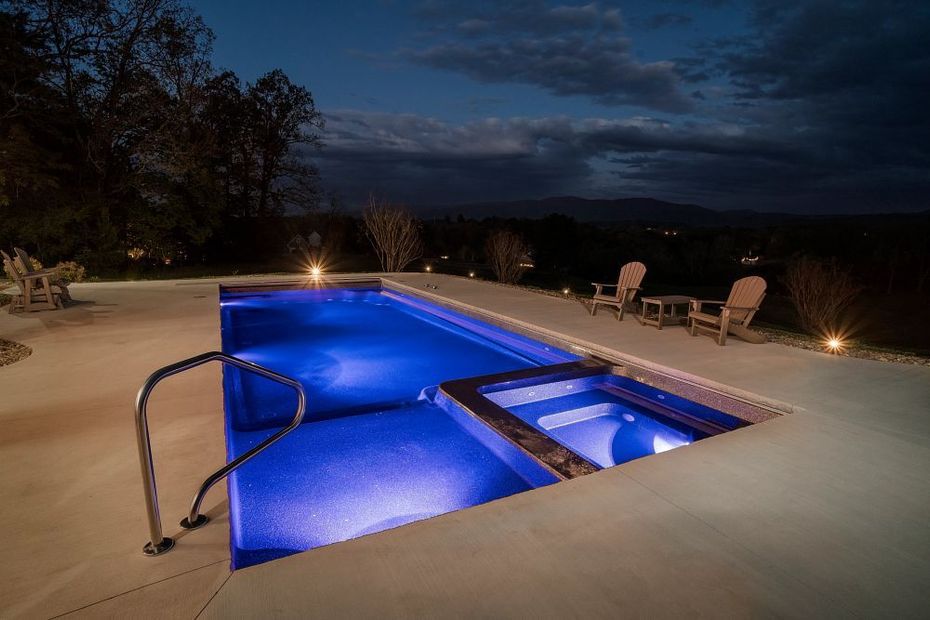 Blue-lit backyard pool at dusk with a spa and lounge chairs beside it