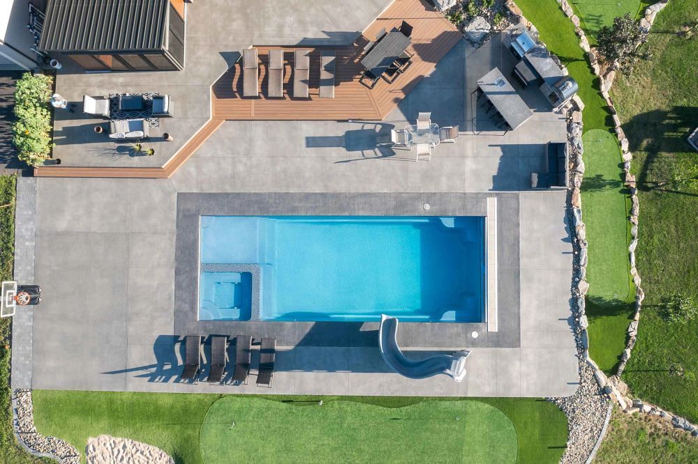 Aerial view of a backyard with a rectangular swimming pool, patio seating, and green lawn.