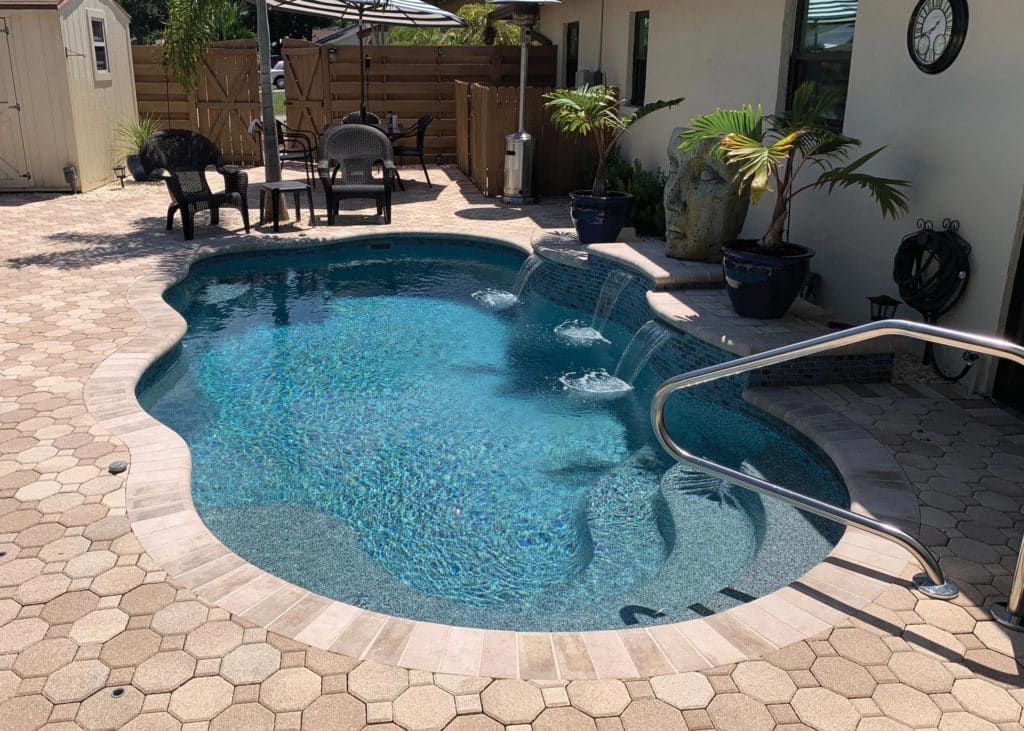 Backyard swimming pool with blue water, stone pavers, patio furniture, and potted plants in shade