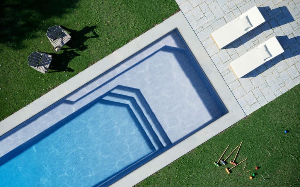 Aerial view of a backyard pool beside green lawn, lounge chairs, and patio furniture.