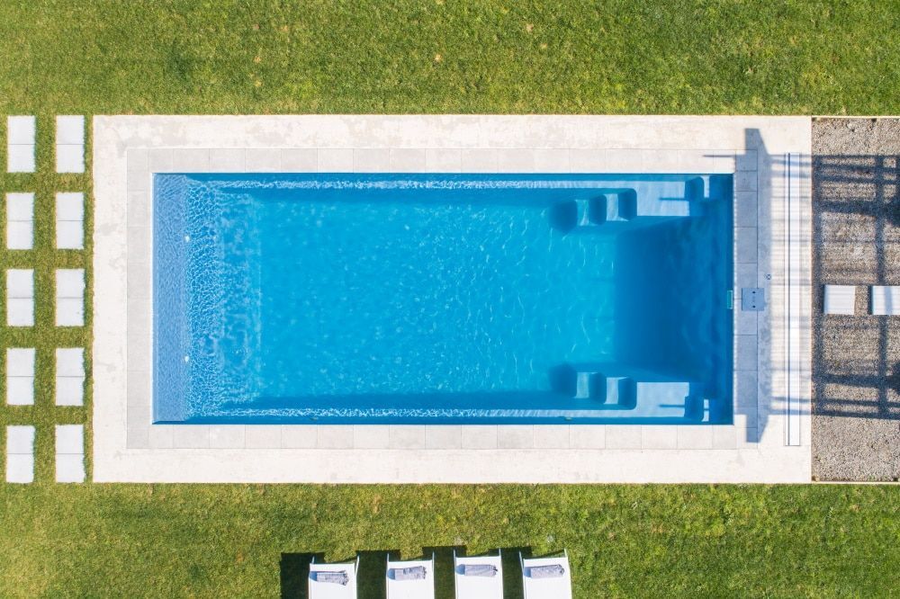 Overhead view of a rectangular blue swimming pool bordered by white decking and green grass.