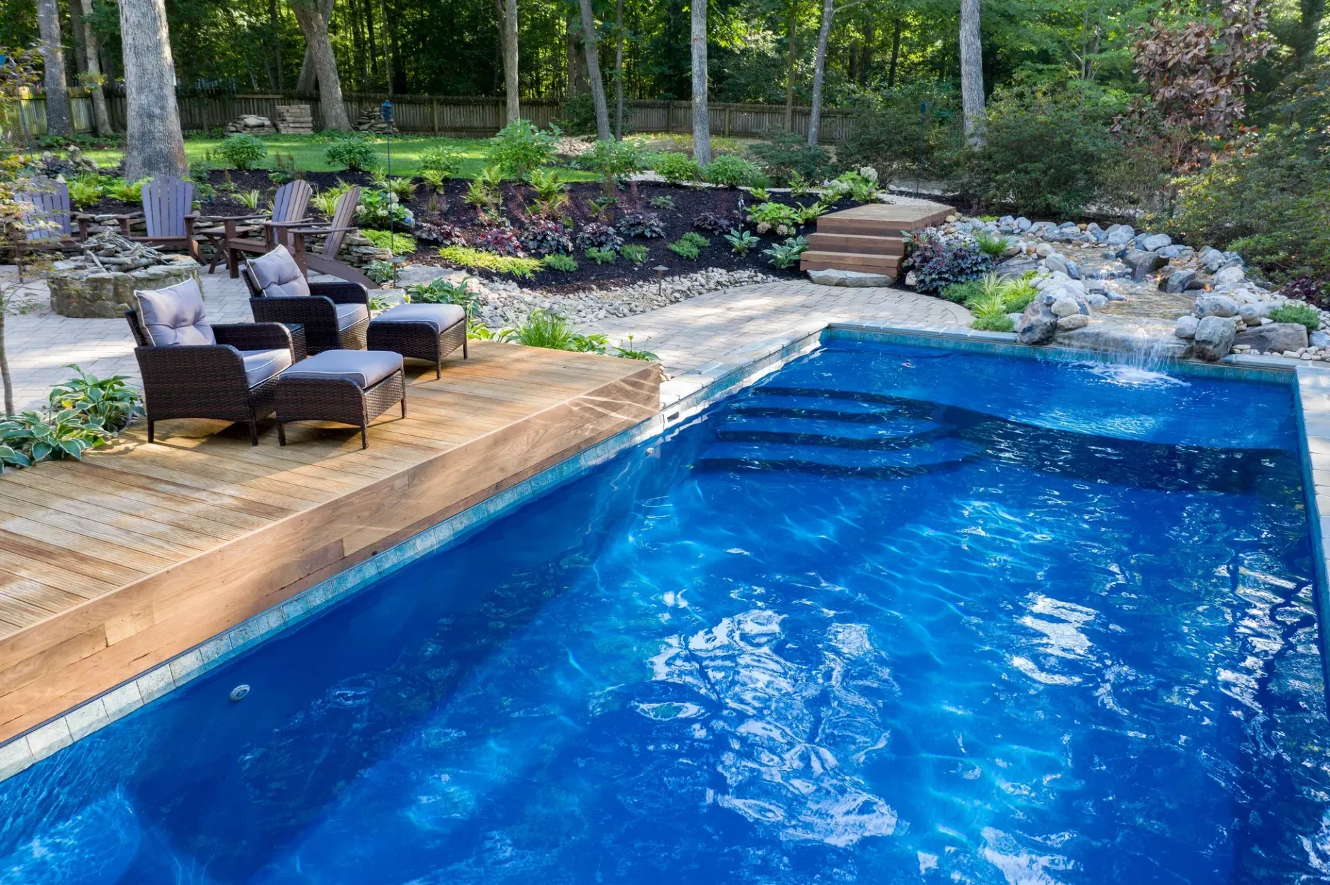 Backyard pool with blue water, wooden deck, lounge chairs, and stone steps surrounded by greenery