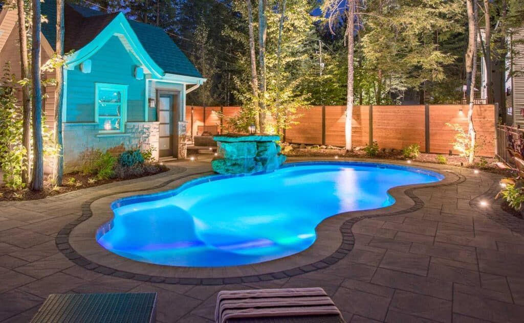 Blue-lit backyard swimming pool beside a small turquoise pool house at night