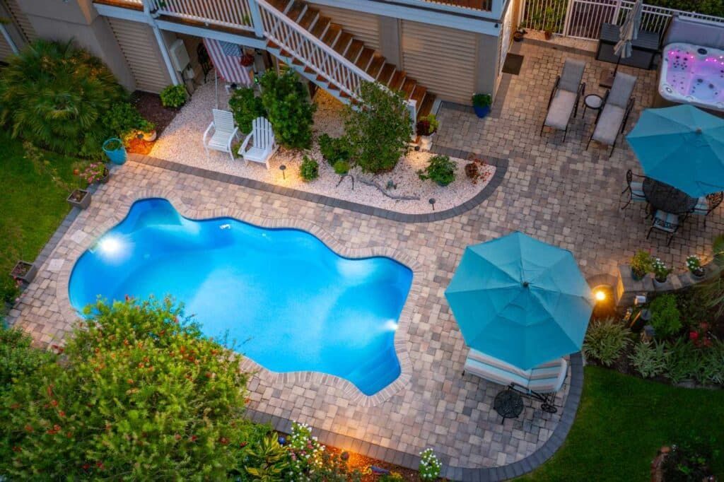 Backyard pool courtyard with blue umbrella, lounge chairs, and glowing pool lights at dusk