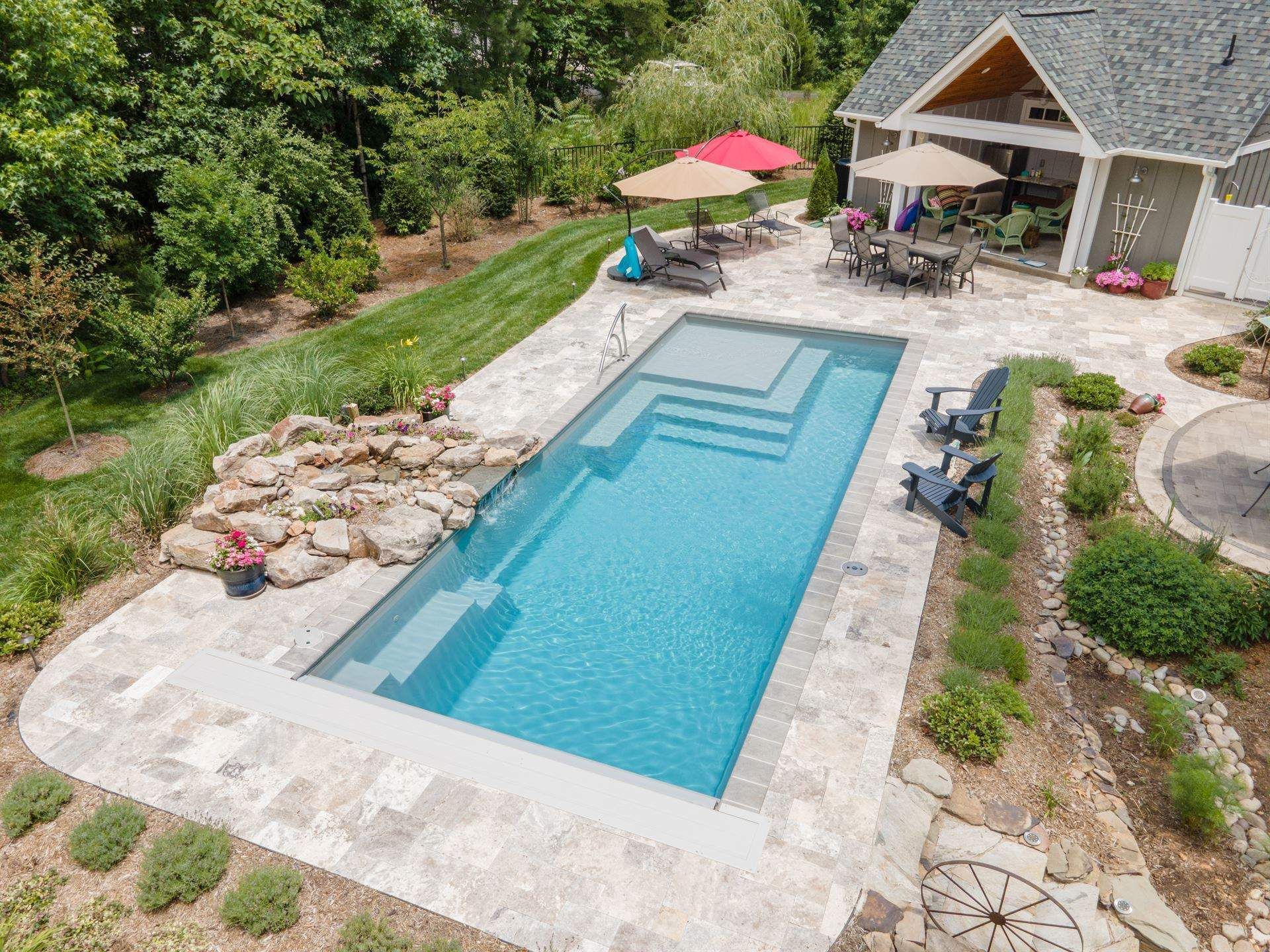 Backyard with a rectangular pool, lounge chairs, patio, and red umbrella beside a house and garden.