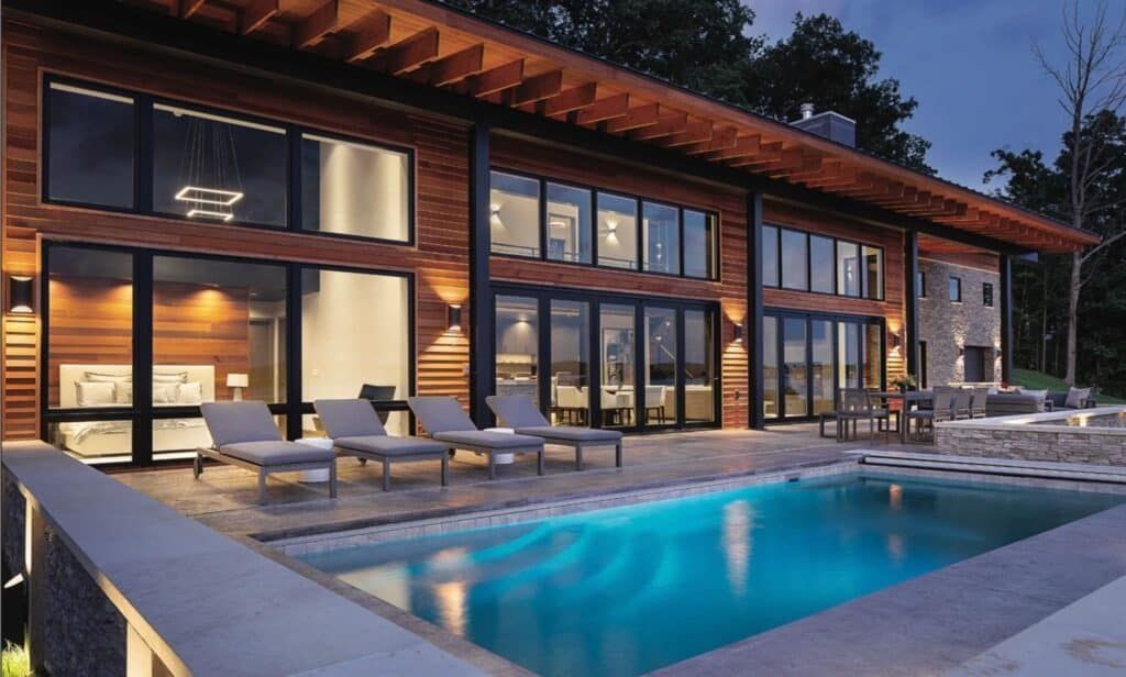 Modern glass house at dusk with a lit patio and a turquoise swimming pool