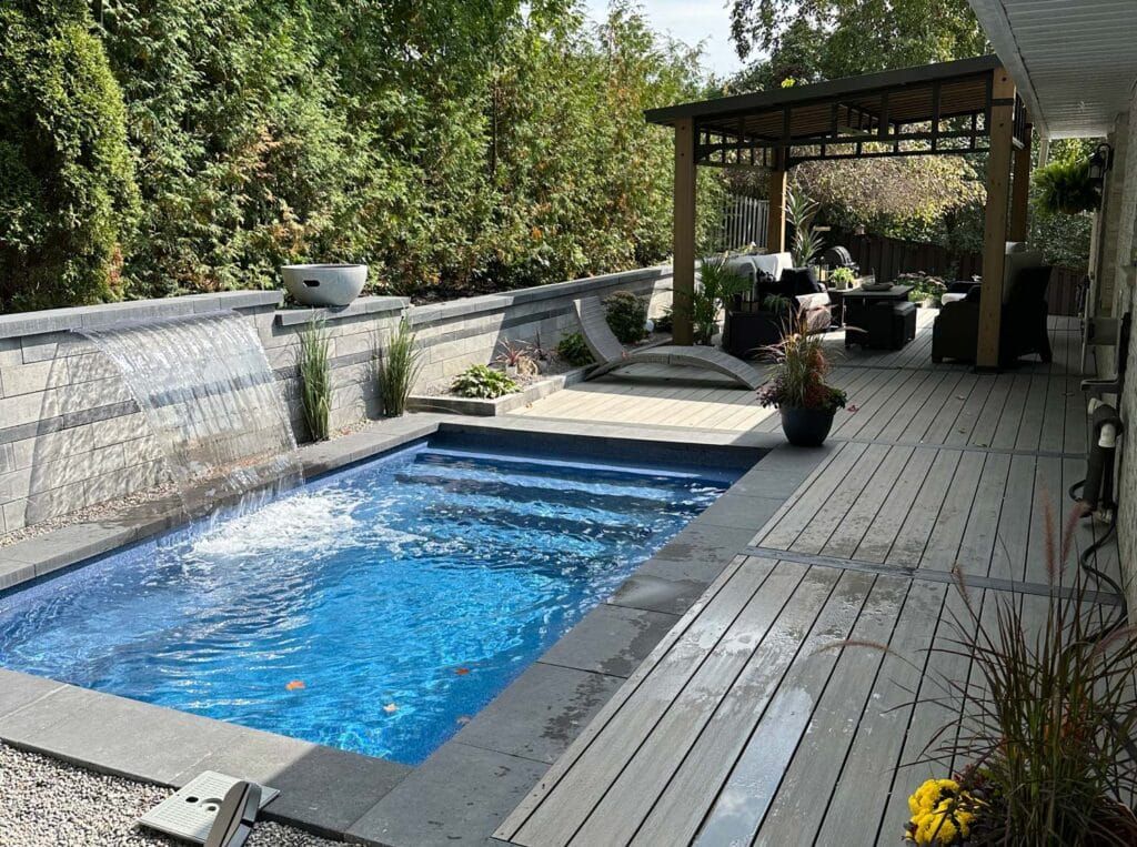 Backyard deck with a small rectangular pool, lounge chairs, and a covered patio beside greenery