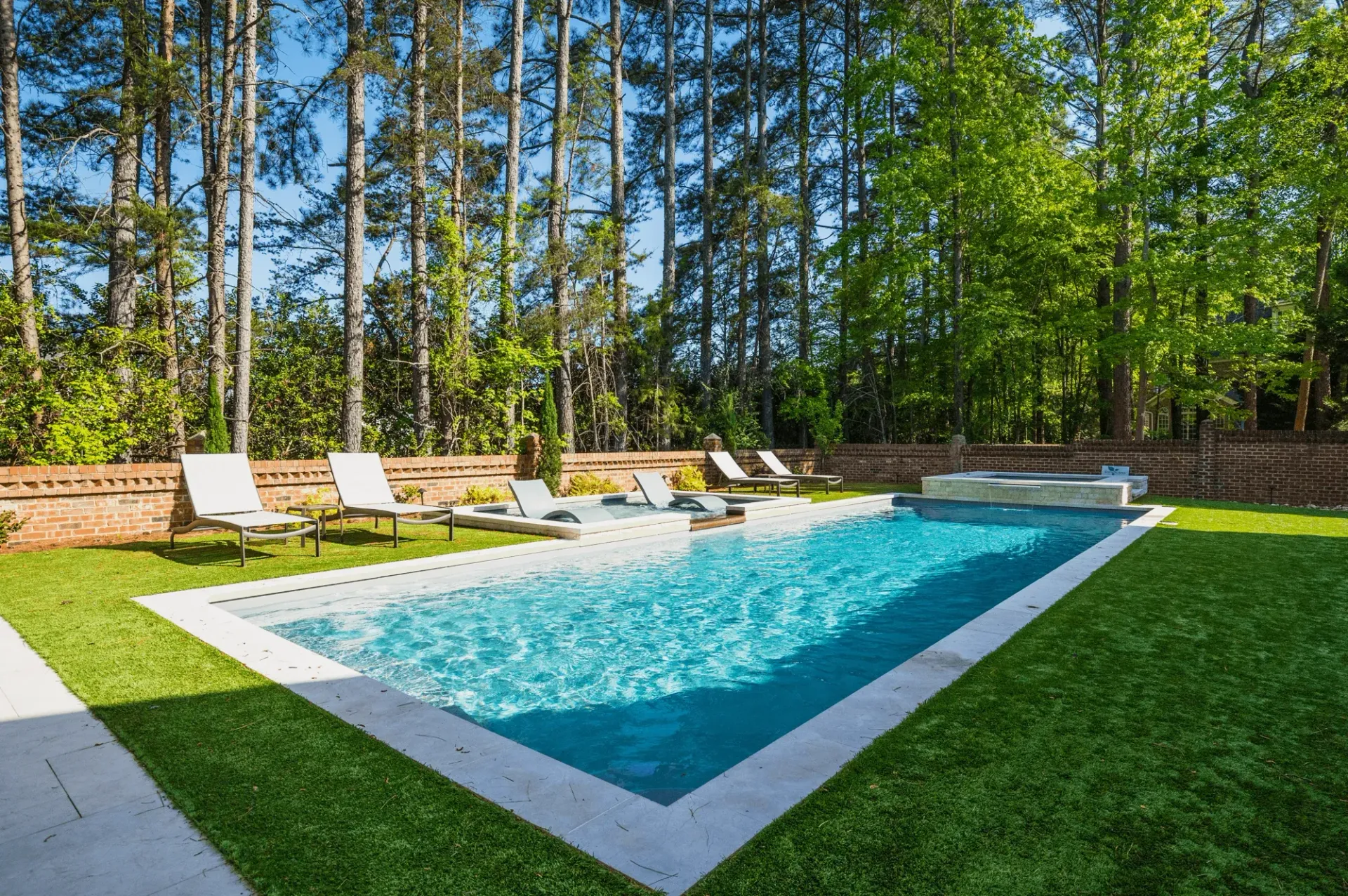 Rectangular backyard pool with white lounge chairs, green lawn, and tall trees around it