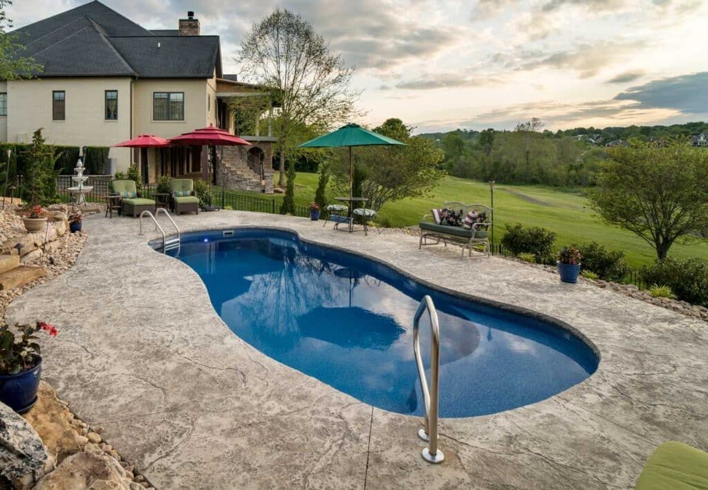Backyard with a kidney-shaped blue swimming pool beside a house and grassy countryside view.