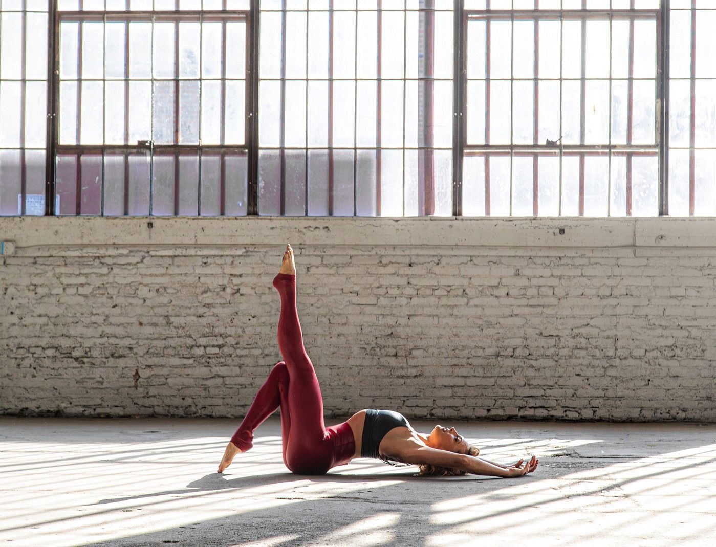 Person practicing yoga on a sunlit floor, lying on their back with one leg raised near large windows.