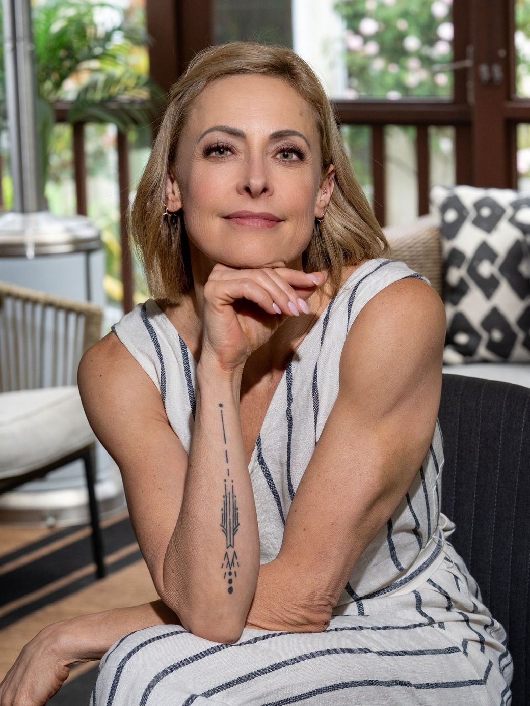 Woman in a sleeveless striped dress posing indoors by a window, resting her chin on her hand.