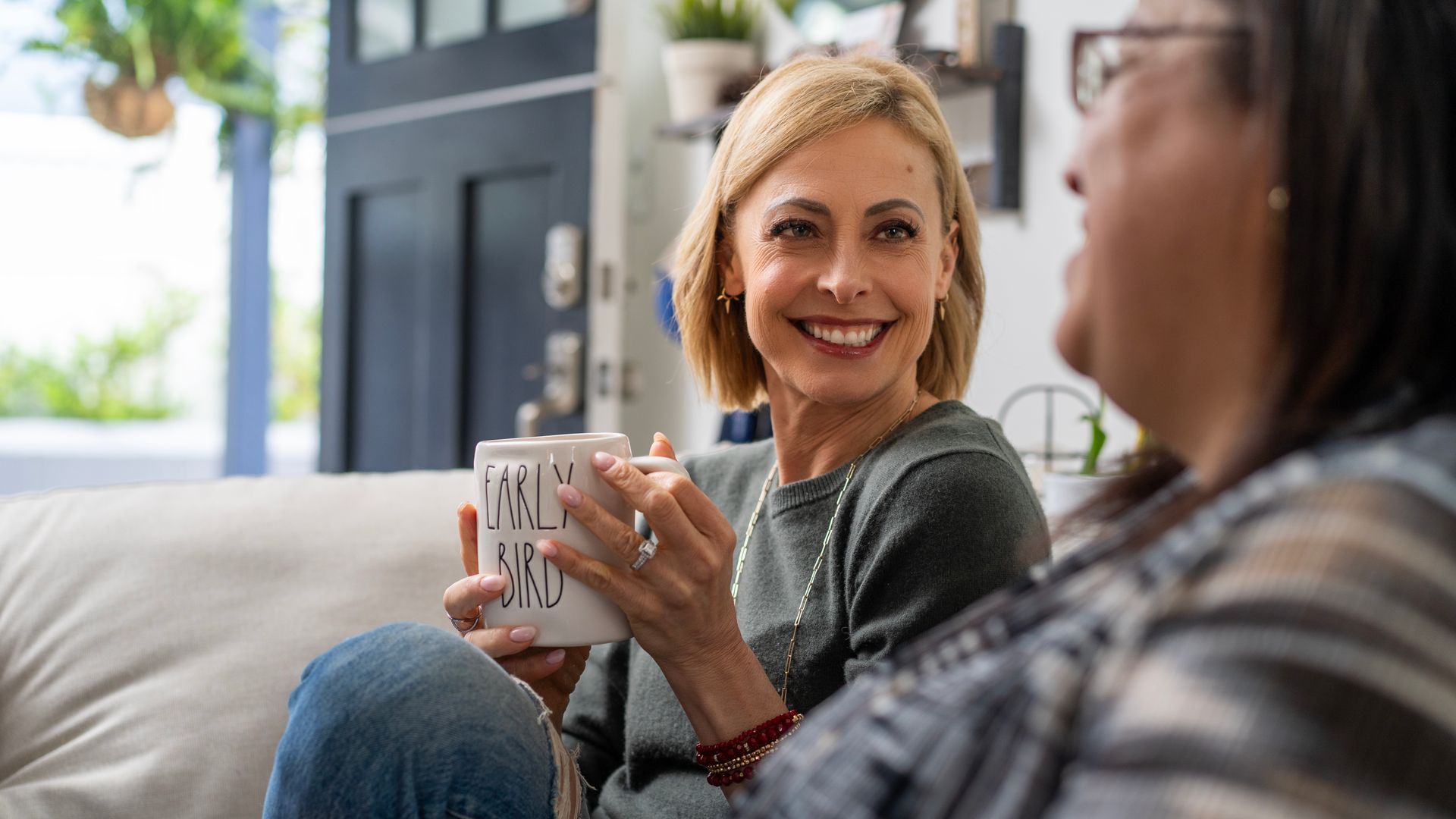 Two people chatting on a couch, one smiling and holding a mug in a bright living room.