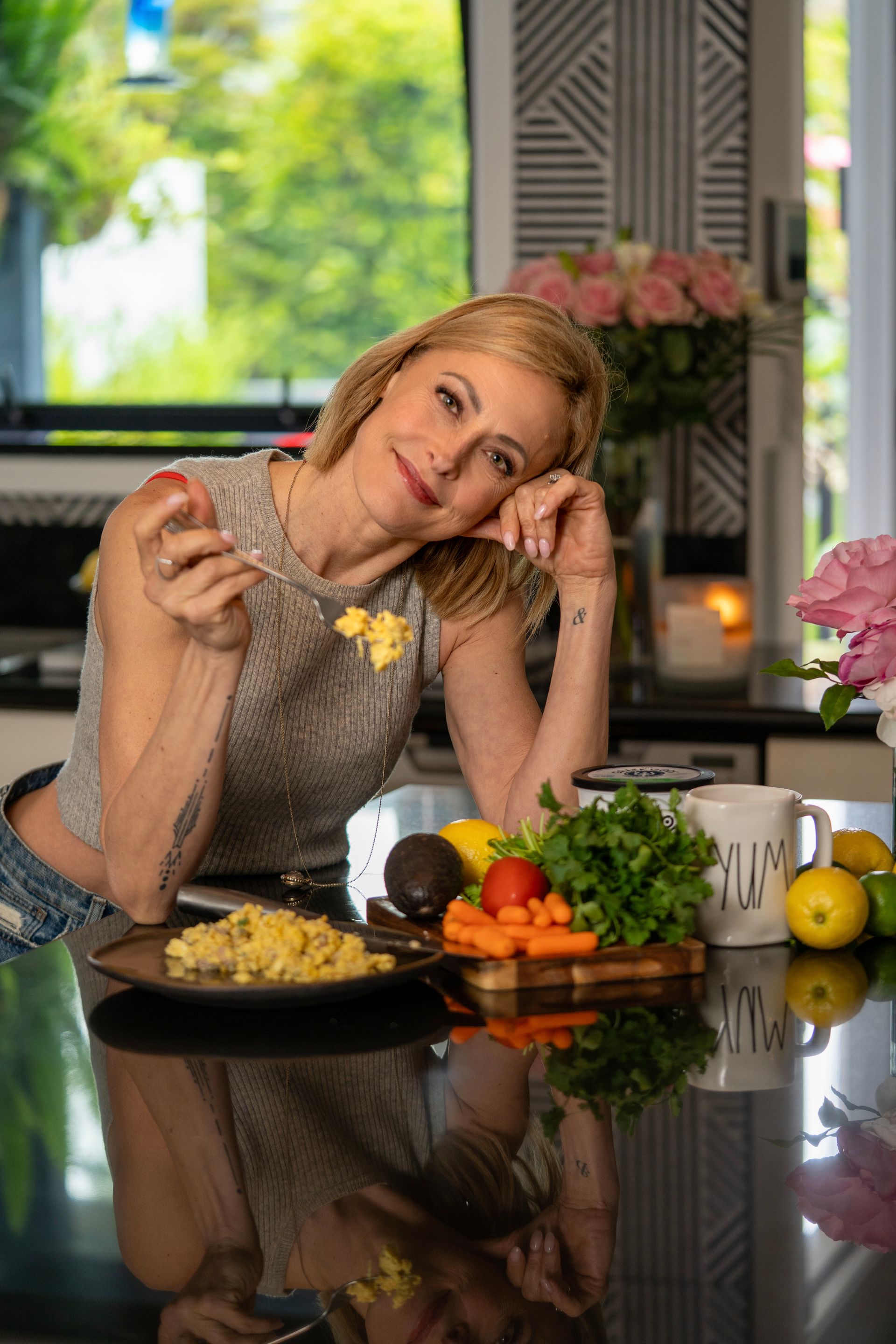 Woman smiling at a kitchen counter with fresh produce, holding a spoon and posing beside prepared food.