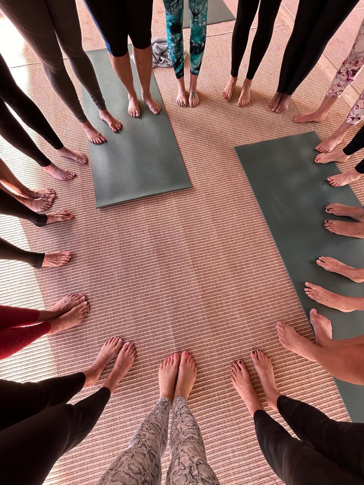 People standing in a circle with hands together over a patterned floor, with yoga mats nearby