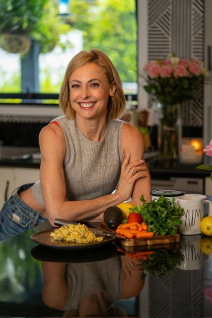 Woman smiling in a kitchen beside fresh vegetables and pasta on a black countertop