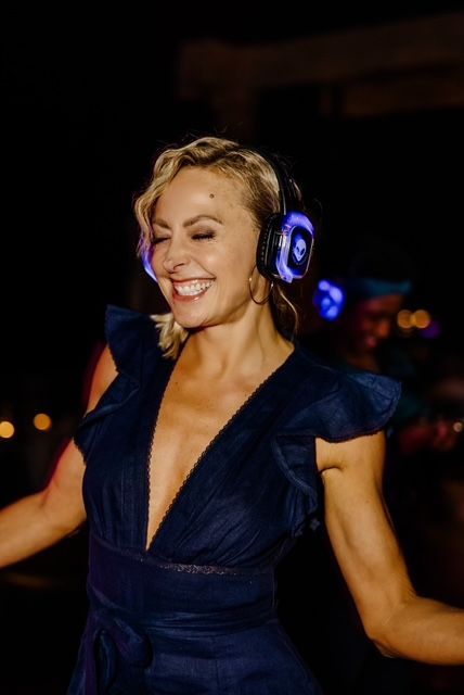 Smiling person in a dark blue dress dancing with glowing headphones on in a dim venue