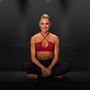 Woman seated cross-legged in a red sports bra against a dark background, smiling at the camera