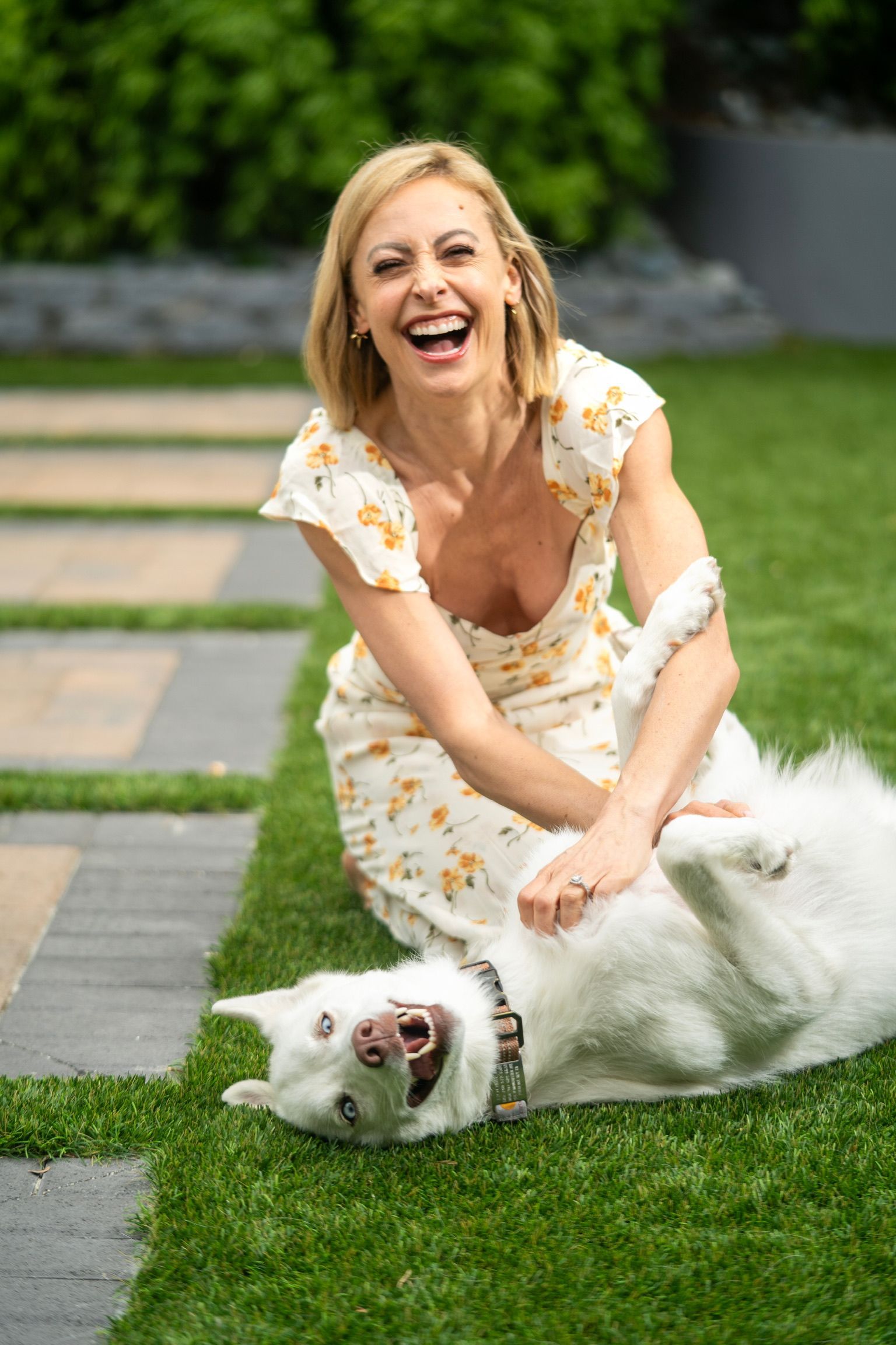 Woman laughing while petting a white dog on a lawn near a stone path