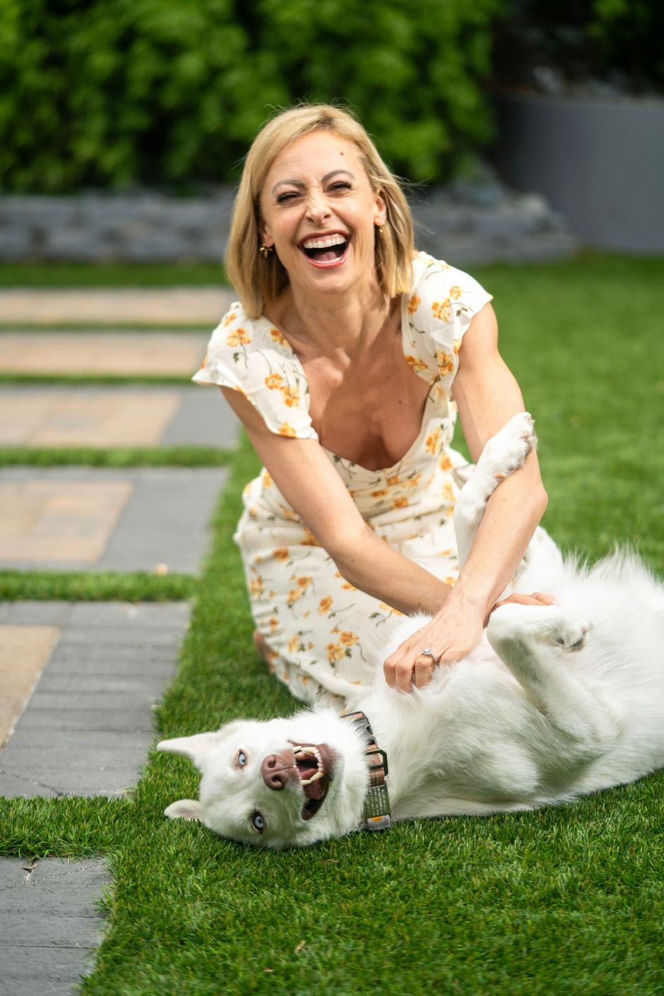 Woman laughing while petting a white dog on a lawn near a stone path