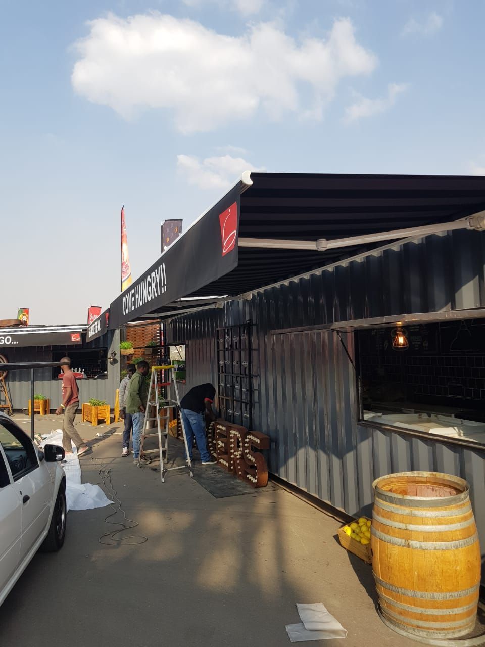 A shipping container stall with a black awning and wooden barrel in a construction area where people are working.
