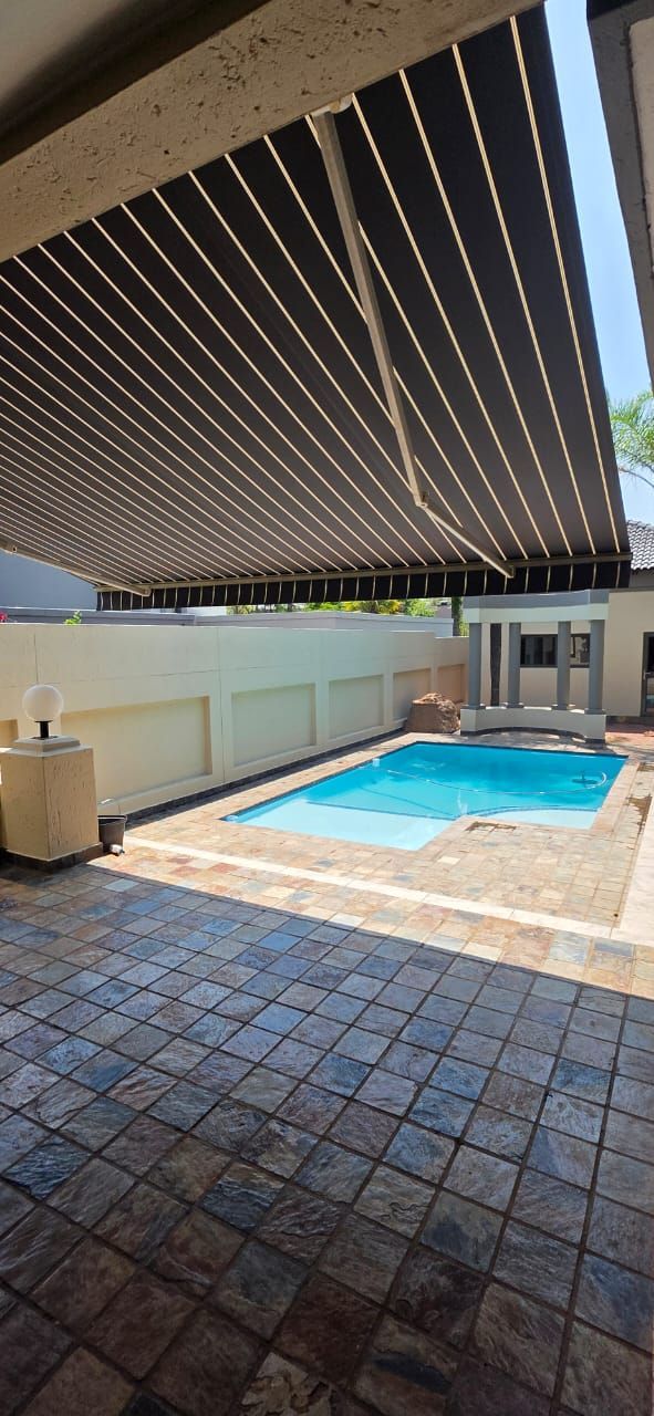 A patio with patterned stone pavers under a dark retractable awning, featuring a small rectangular pool in the background.
