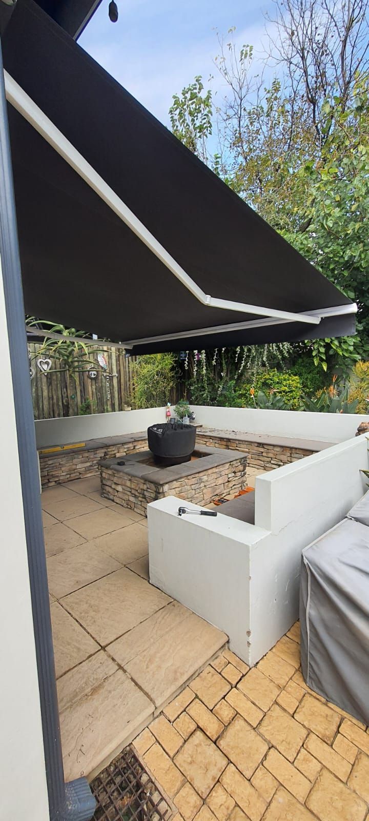 A patio area with a stone fire pit, built-in benches, light-colored pavers, and a large black retractable awning overhead.