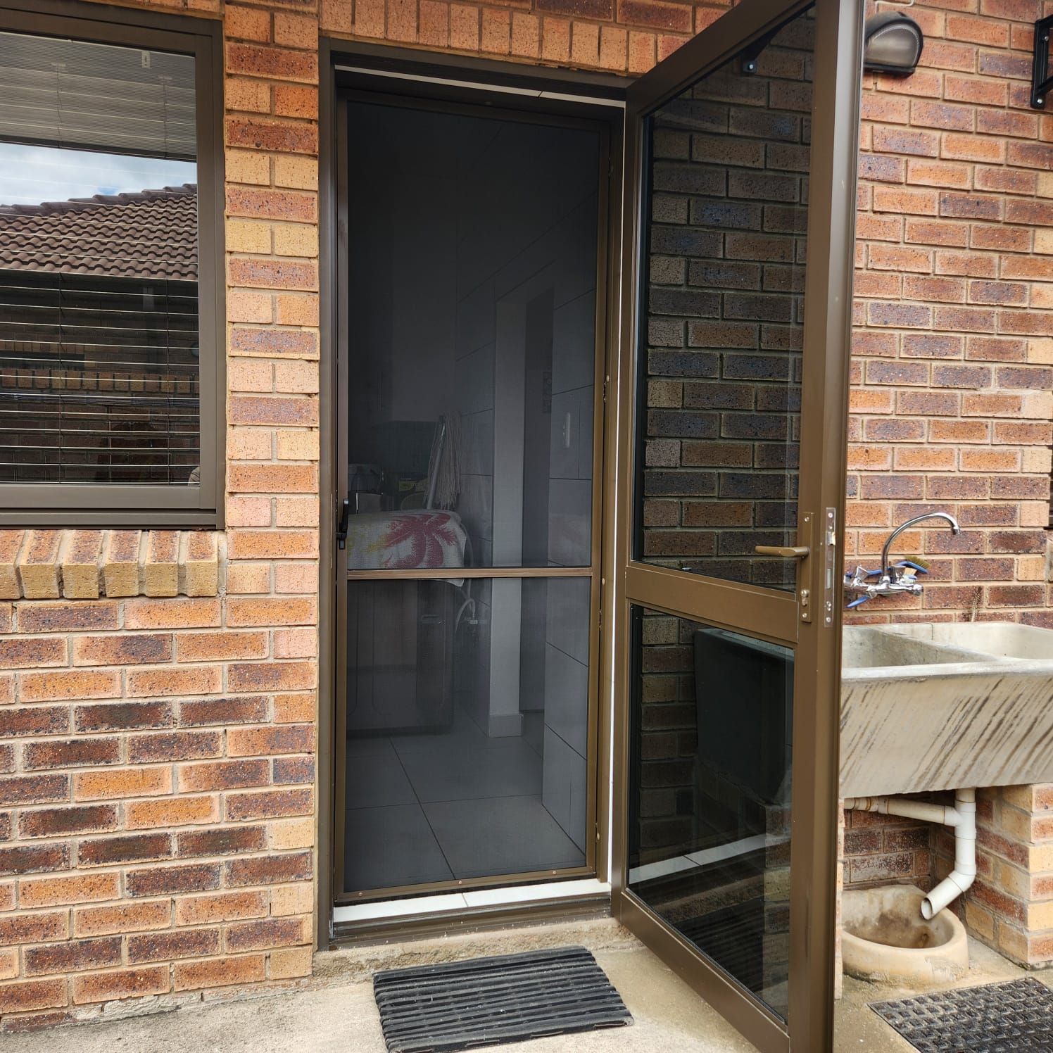 A partially open bronze wildlife screen door on a brick wall with a nearby outdoor sink.