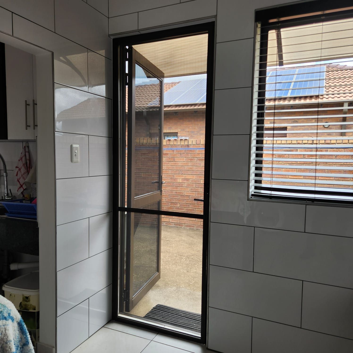 A white tiled kitchen area featuring a dark-framed wildlife screen door open and a window with blinds.