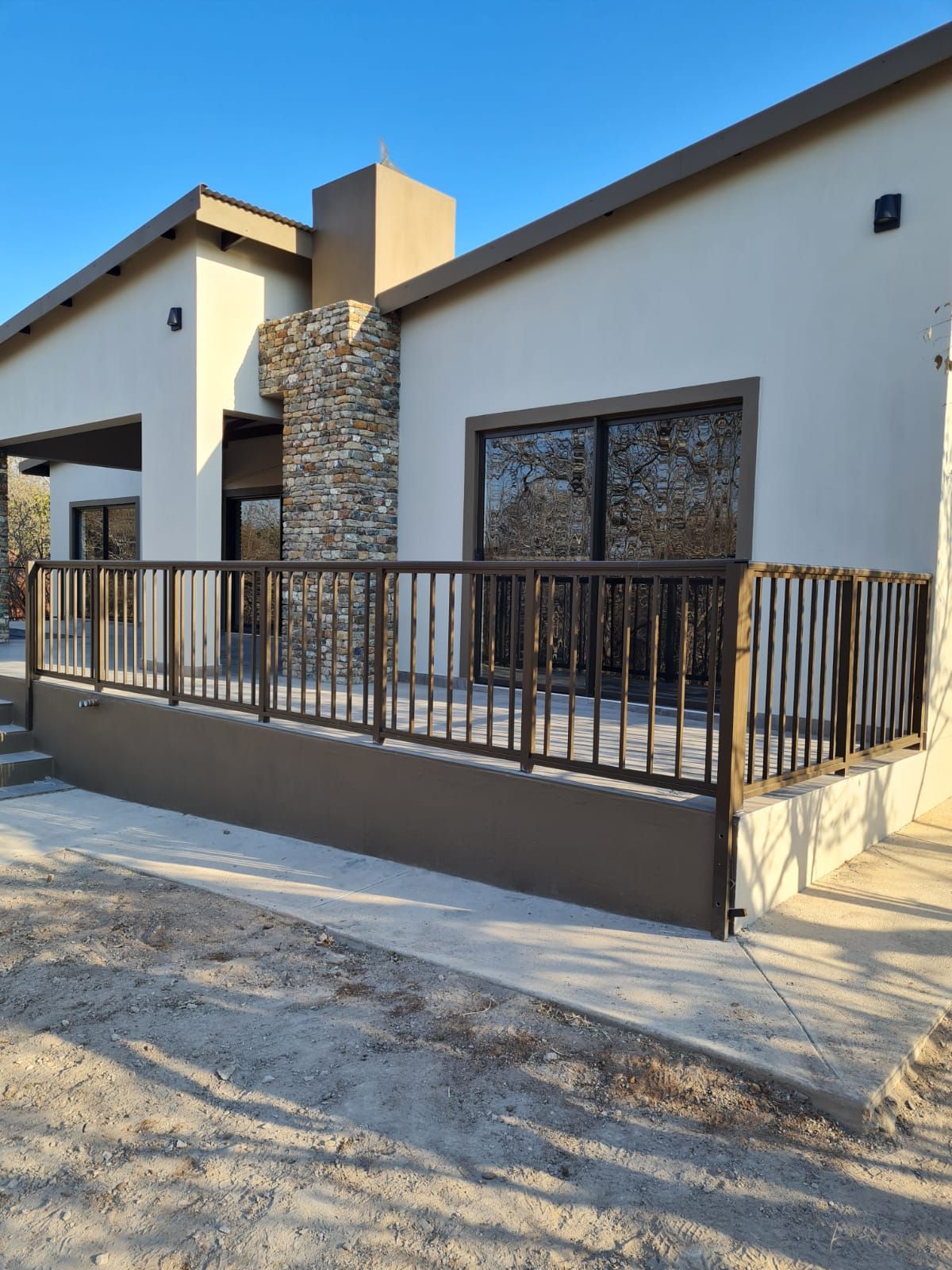 A modern house with a stone chimney, tan walls, and a deck with a dark metal railing under a clear blue sky.
