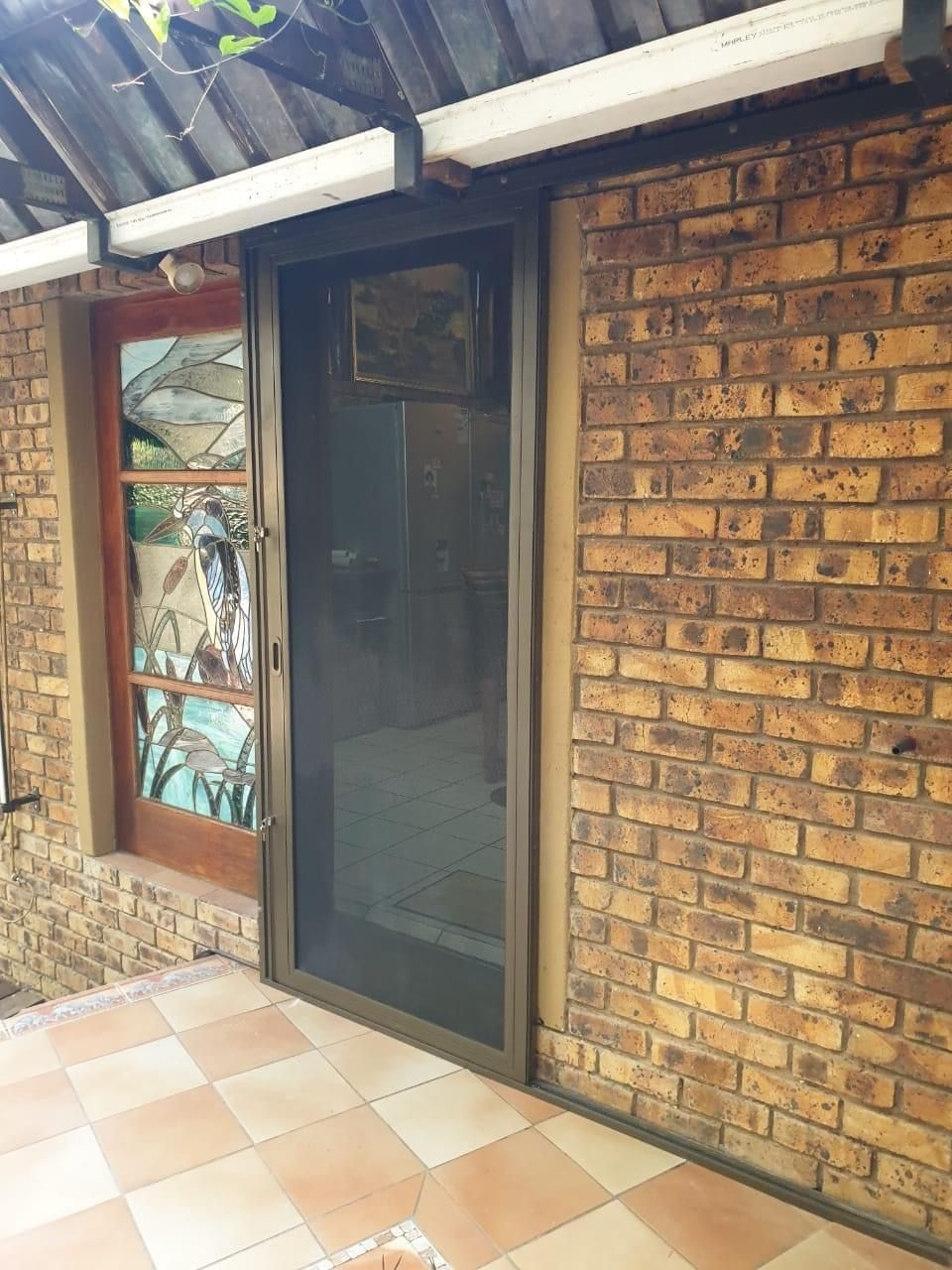 A screen door with a dark mesh panel attached to a brown brick wall beside a stained-glass door on a tiled patio.
