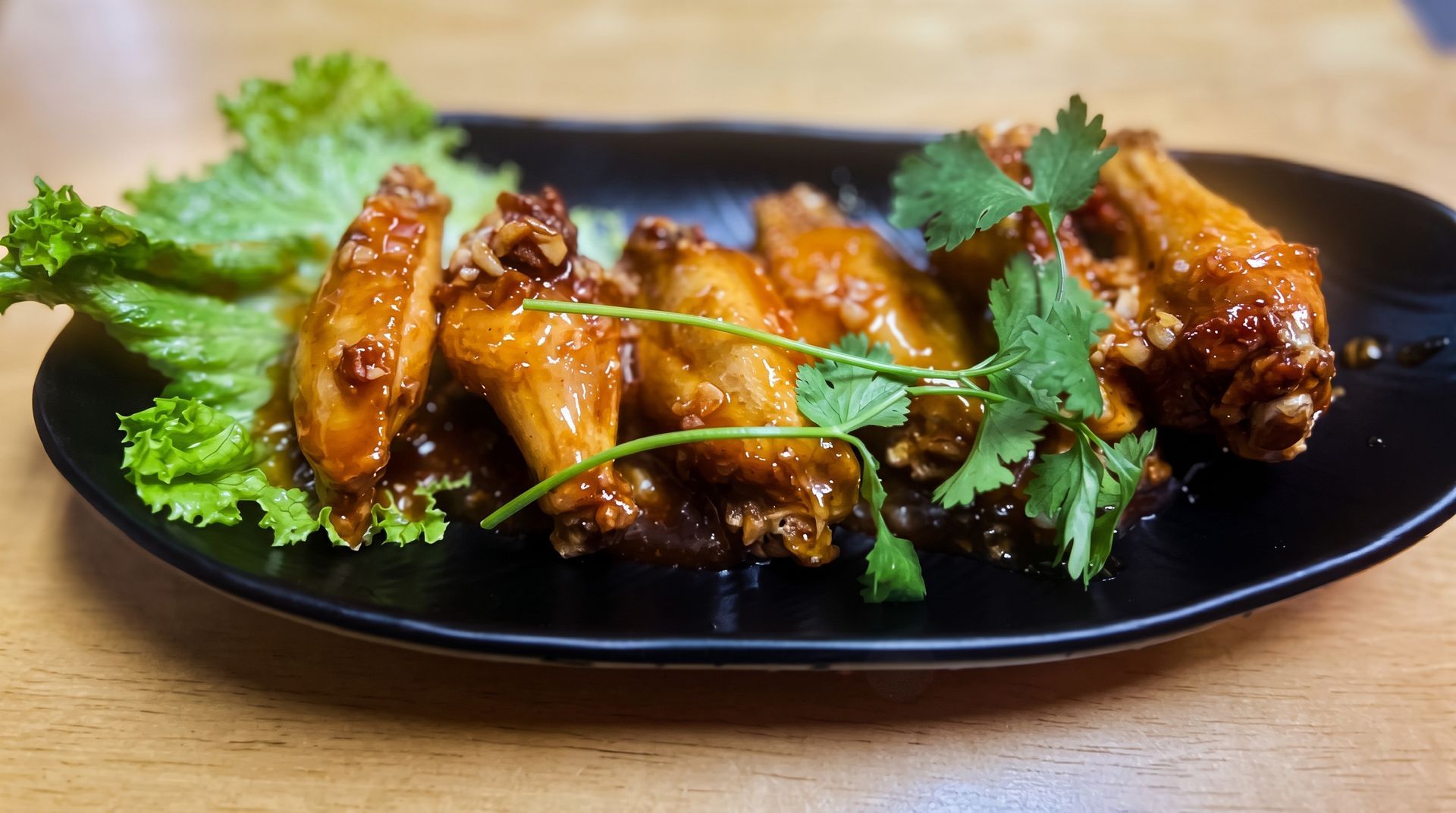 Glazed chicken wings on a black plate, garnished with lettuce and cilantro on a wooden table