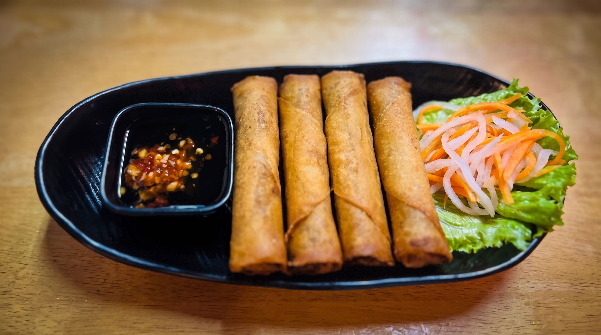 Fried spring rolls on a black platter with dipping sauce and a small salad garnish