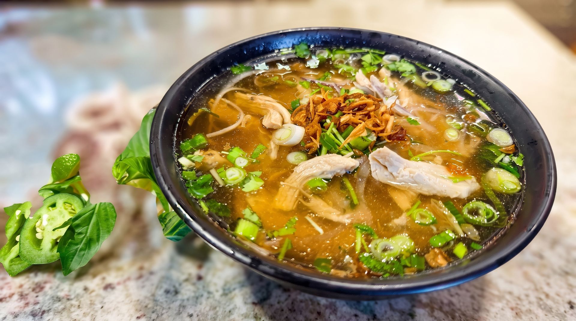 Black bowl of noodle soup topped with herbs and meat on a table, with lime and greens beside it