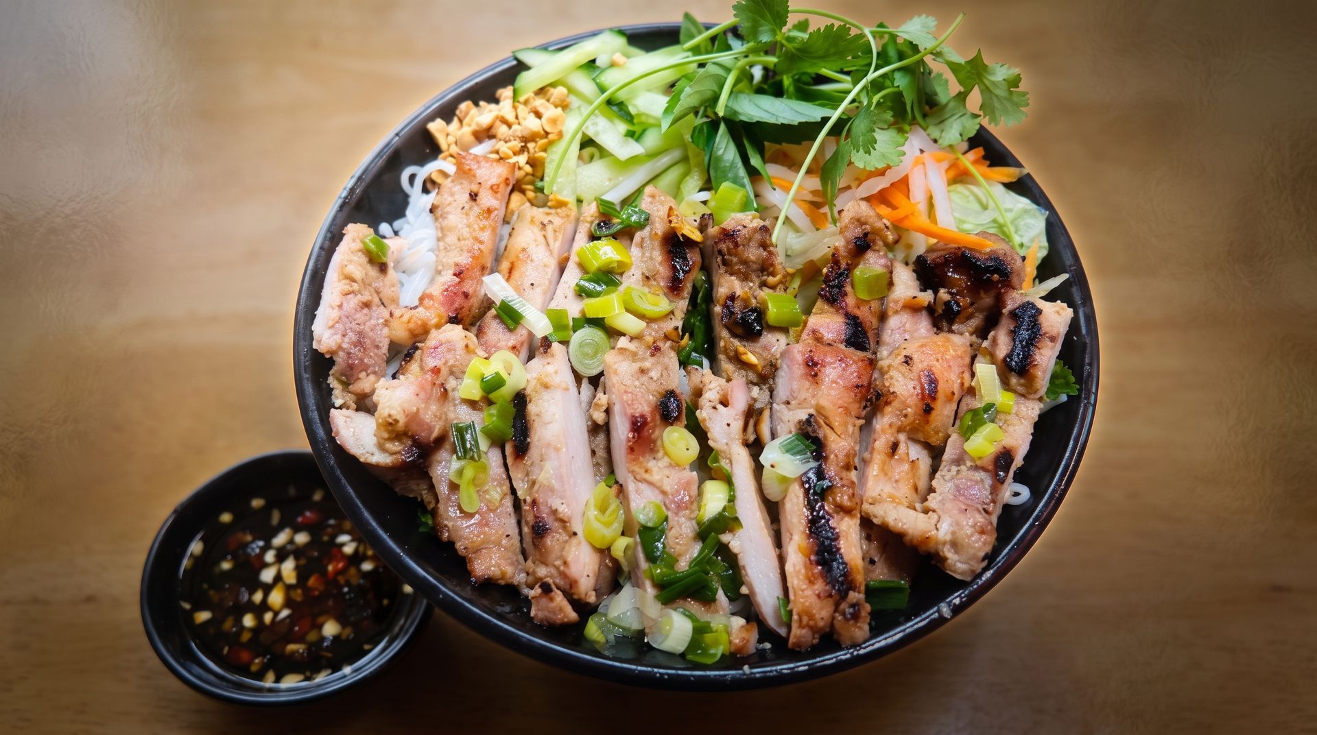 Grilled meat bowl with rice, greens, and vegetables, served with dipping sauce on a wooden table