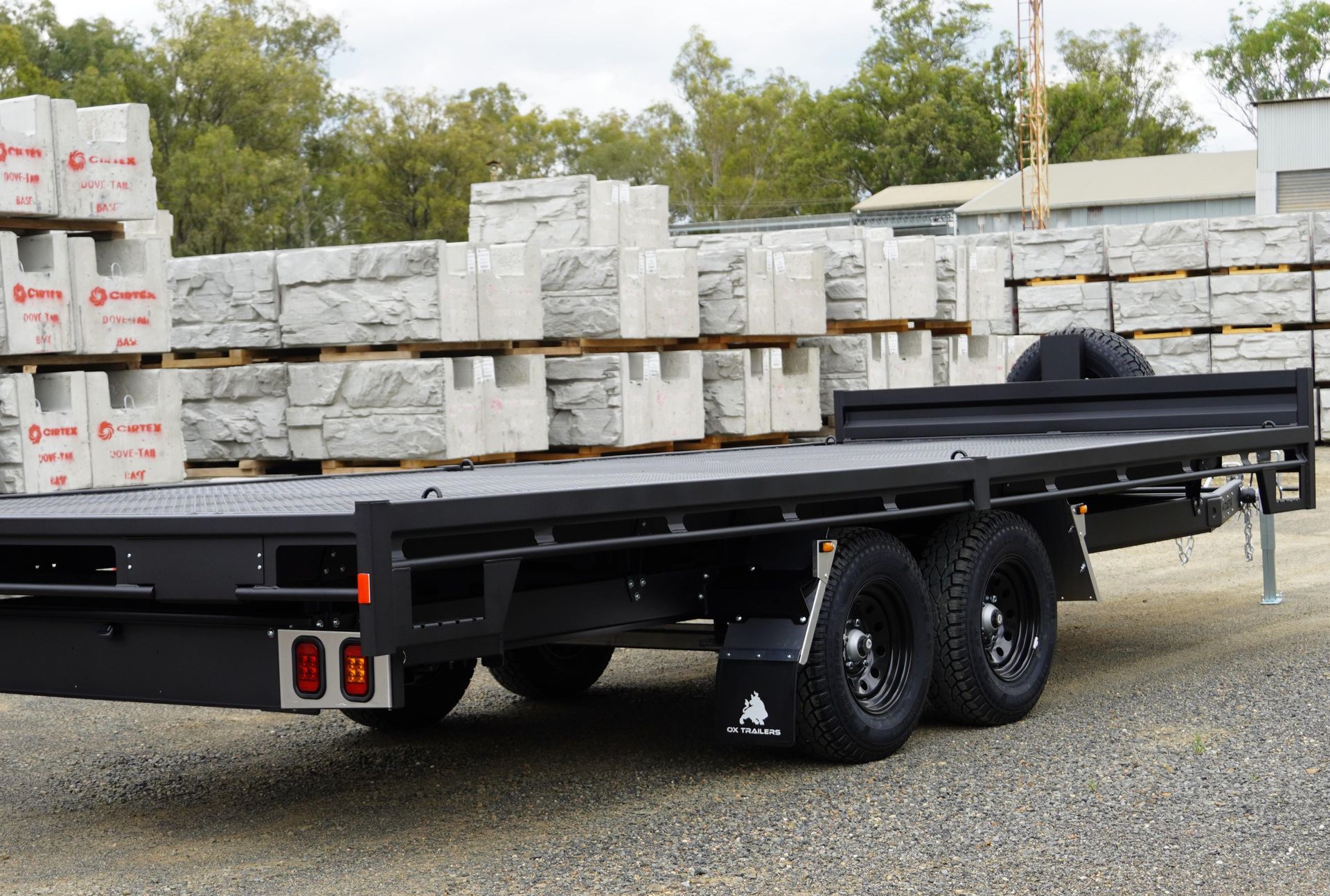 A 6m x 2.4m Black Flat Top Trailer with a headboard, placed between large concrete blocks, without dropsides.