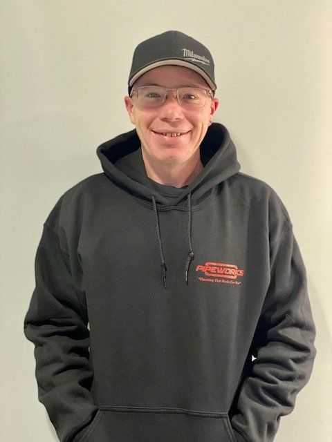 Man in black hoodie and hat, wearing glasses, smiling in front of a light gray wall.