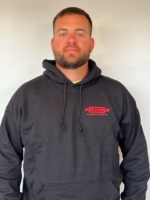 A man with a beard wearing a black hoodie with a logo stands against a white wall. He is looking at the camera.