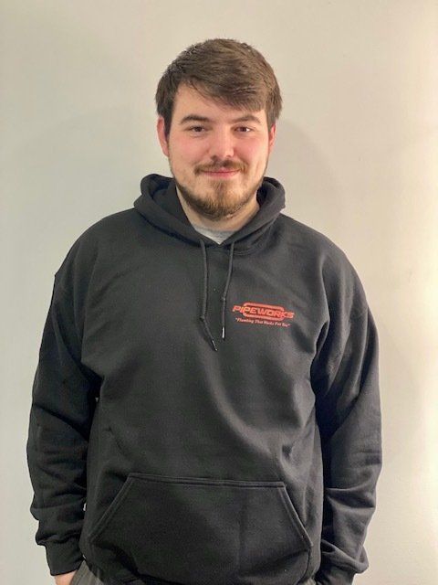 A young man with a beard in a black hoodie stands in front of a white wall.