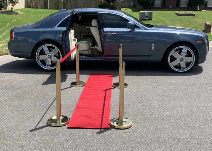 Blue luxury car with open door beside a red carpet walkway and stanchions on a street