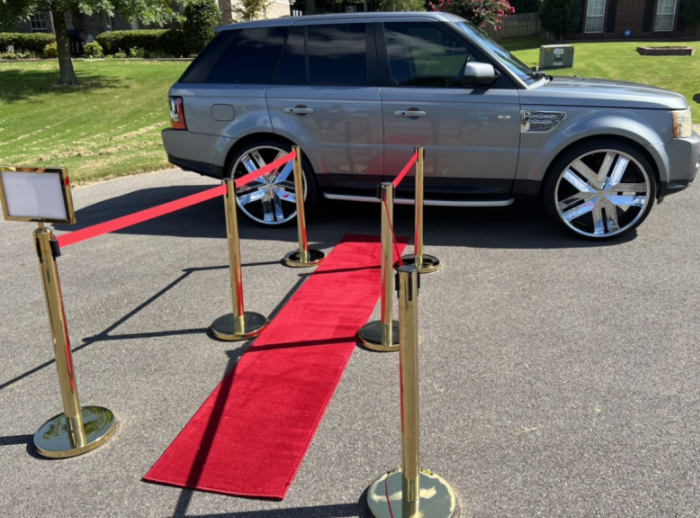Gray SUV parked beside a red carpet walkway with gold stanchions on a paved road.