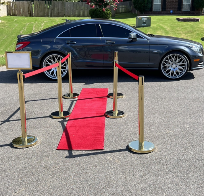 Black luxury sedan beside a red carpet and gold stanchions on a driveway
