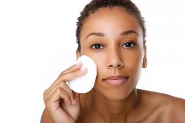 Woman applying a white facial pad to her cheek against a plain white background