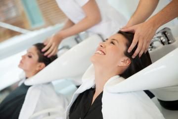 Two women at salon sinks getting their hair washed by stylists, smiling and reclining in white chairs