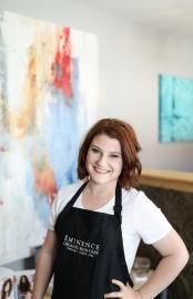 Smiling woman in a black apron stands in an art studio beside colorful abstract paintings